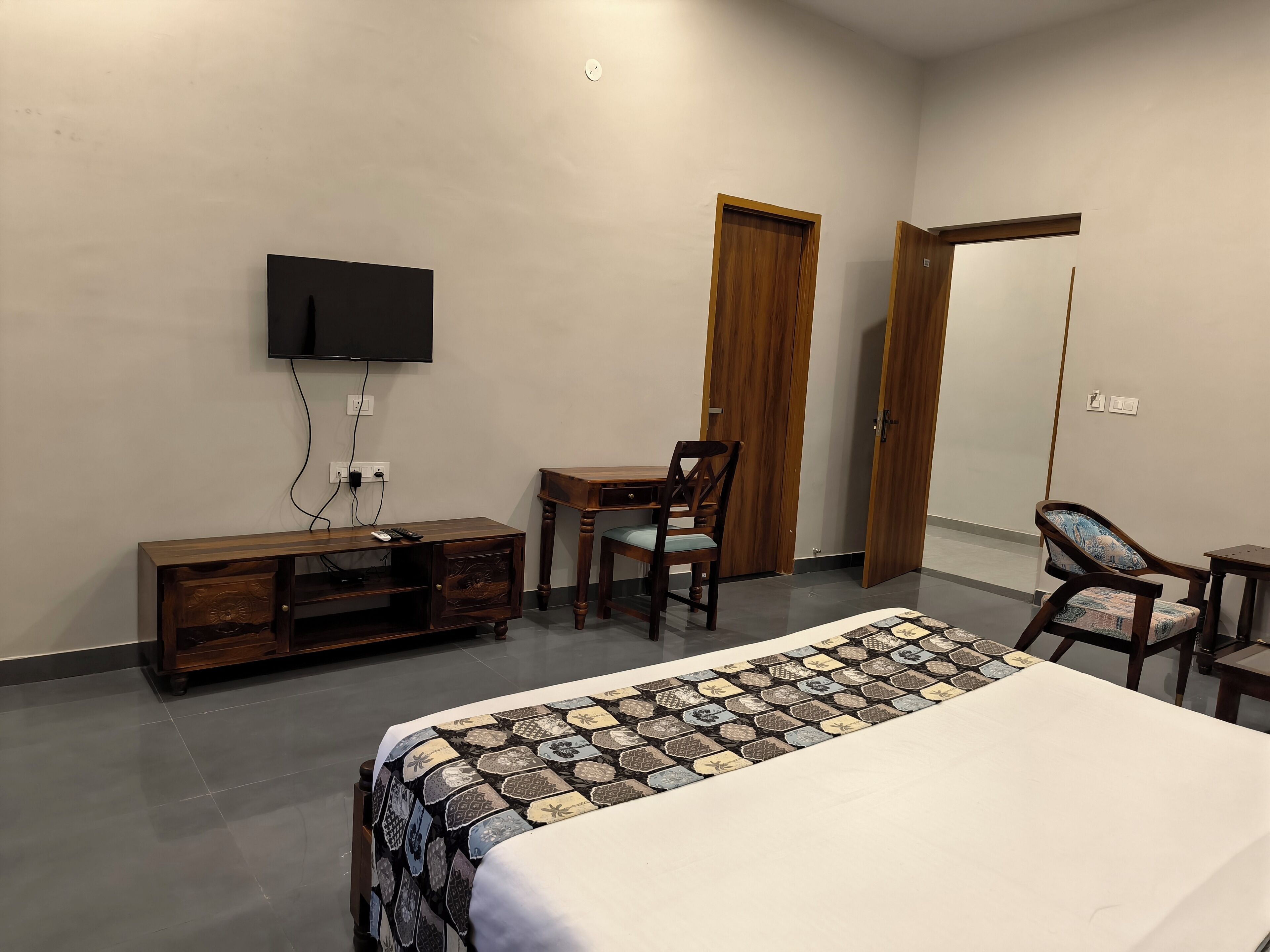 A hotel room with a bed, a desk, a chair, a TV mounted on the wall, and two wooden doors.