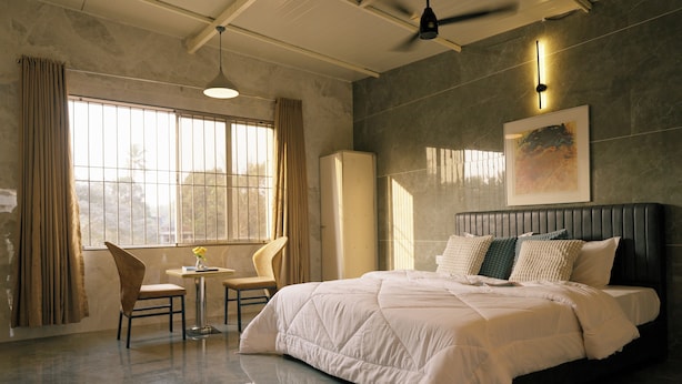 A bedroom with a large bed, a chair, a small table, and a window with curtains. - SHREE LODGING & BOARDING (Vasai)