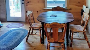 A cozy wooden cabin with a round dining table and four chairs, a ceiling fan, and a window with a view of trees. - Kettlestone (Stanton)