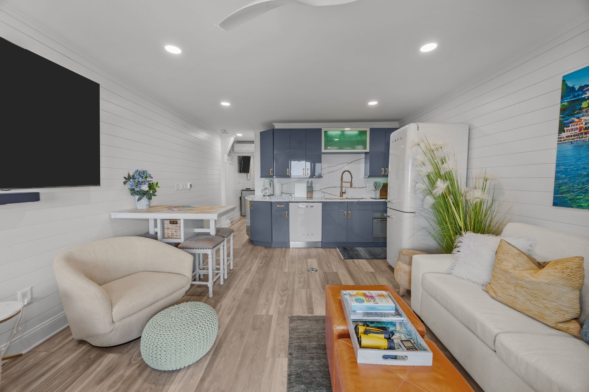 A modern living room with a sofa, a coffee table, and a dining area with a table and chairs. A kitchen with grey cabinets and a refrigerator is visible in the background.