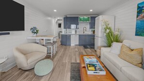 Uma sala de estar moderna com sofá, mesa de centro e área de jantar com mesa e cadeiras. Uma cozinha com armários cinza e uma geladeira é visível ao fundo.
