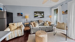 Un salon comprenant un canapé, un fauteuil et une table basse. On y trouve également un ventilateur de plafond, une lampe et un tableau accroché au mur.