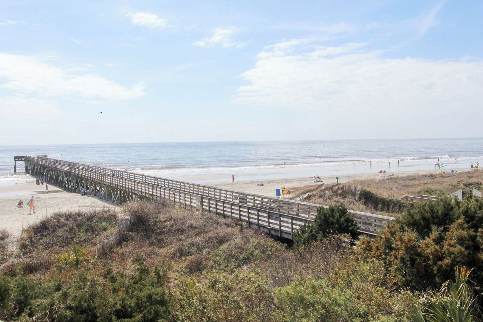 Ein Strand mit einer Holzstege, der sich ins Meer erstreckt, feinem Sand und spärlicher Vegetation.
