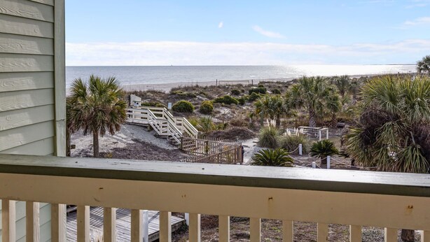 Vue sur la plage depuis un balcon doté d’une balustrade en bois, avec une plage de sable, des palmiers et une clôture en bois.