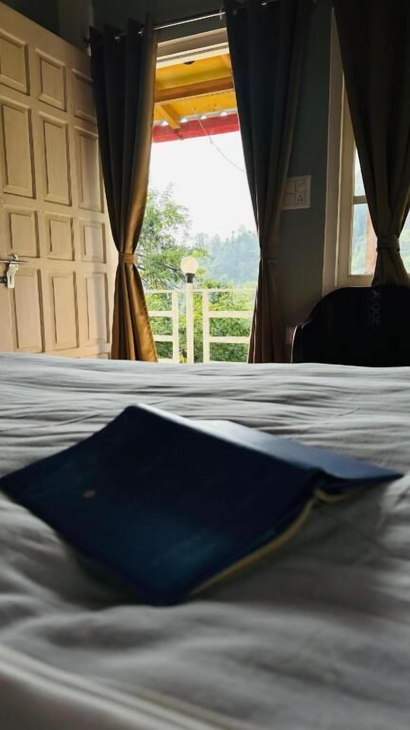 A bed with a blue folder on it, a white door, and a view of greenery outside.
