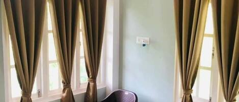 A room with two windows, each with brown curtains tied back, a small table with a chair, and a bed with a blue cover.