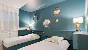 Une chambre d’hôtel avec deux lits simples, une fenêtre avec des rideaux légers, une table de chevet avec une lampe et un miroir fixé au mur.