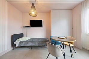 Una habitación moderna con cama, televisor montado en la pared, mesa de comedor con sillas y una ventana con cortinas.