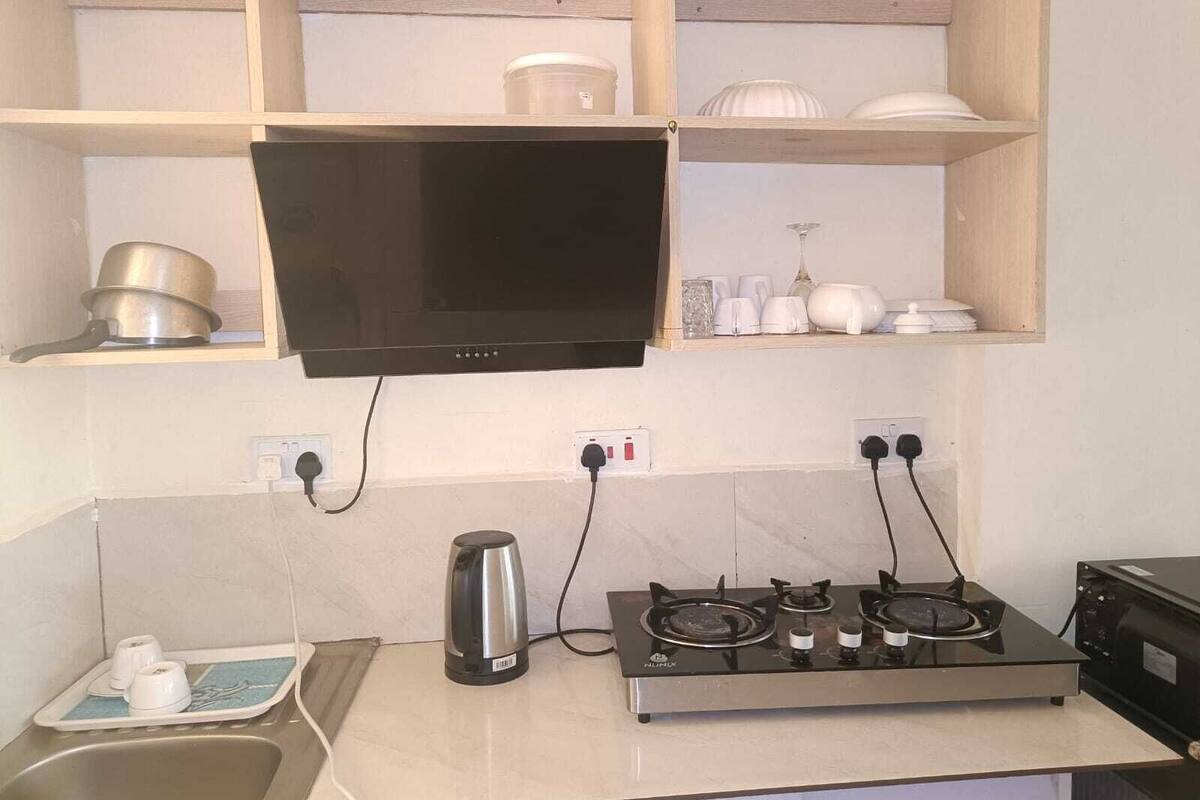 A kitchen with a stove, a kettle, and a microwave. There are shelves above the stove holding dishes and a kettle.