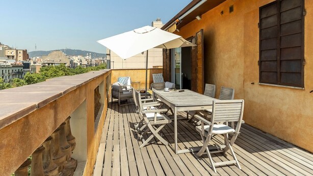 A rooftop terrace with a wooden deck, a table with chairs, a white umbrella, and a city view in the background. - Penthouse with stunning TERRACE / Arco de Triunfo (08009 Barcelona)