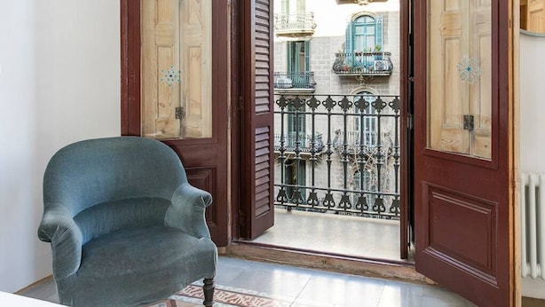 Una habitación con una silla, una puerta de madera y vistas a un balcón con barandillas de hierro y edificios.