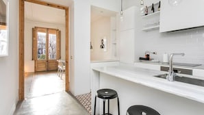 Una cocina moderna con armarios blancos, una isla grande, taburetes de bar, iluminación colgante y vistas a través de una puerta abierta.