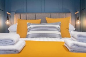 Cama bem arrumada com almofadas amarelas listradas e lisas, cabeceira cinza e duas luminárias fixadas na parede.