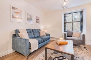 Uma sala de estar moderna com um sofá azul, um sofá cinzento e uma mesa de centro. Há quadros nas paredes e uma janela com persianas.