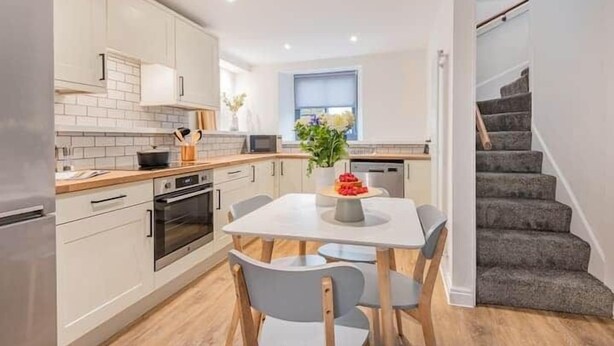 Une cuisine moderne avec des meubles blancs, une table à manger avec des chaises et un escalier menant à l’étage supérieur.
