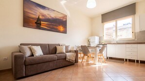 A living room with a brown sofa, a dining table with chairs, a kitchen area with a window, and a painting of a sailboat on the wall. - Apollo Apartment (Palermo)