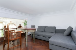 Ein Zimmer mit grauem Sofa, Holzstuhl, rundem Tisch und einer Zimmerpflanze.