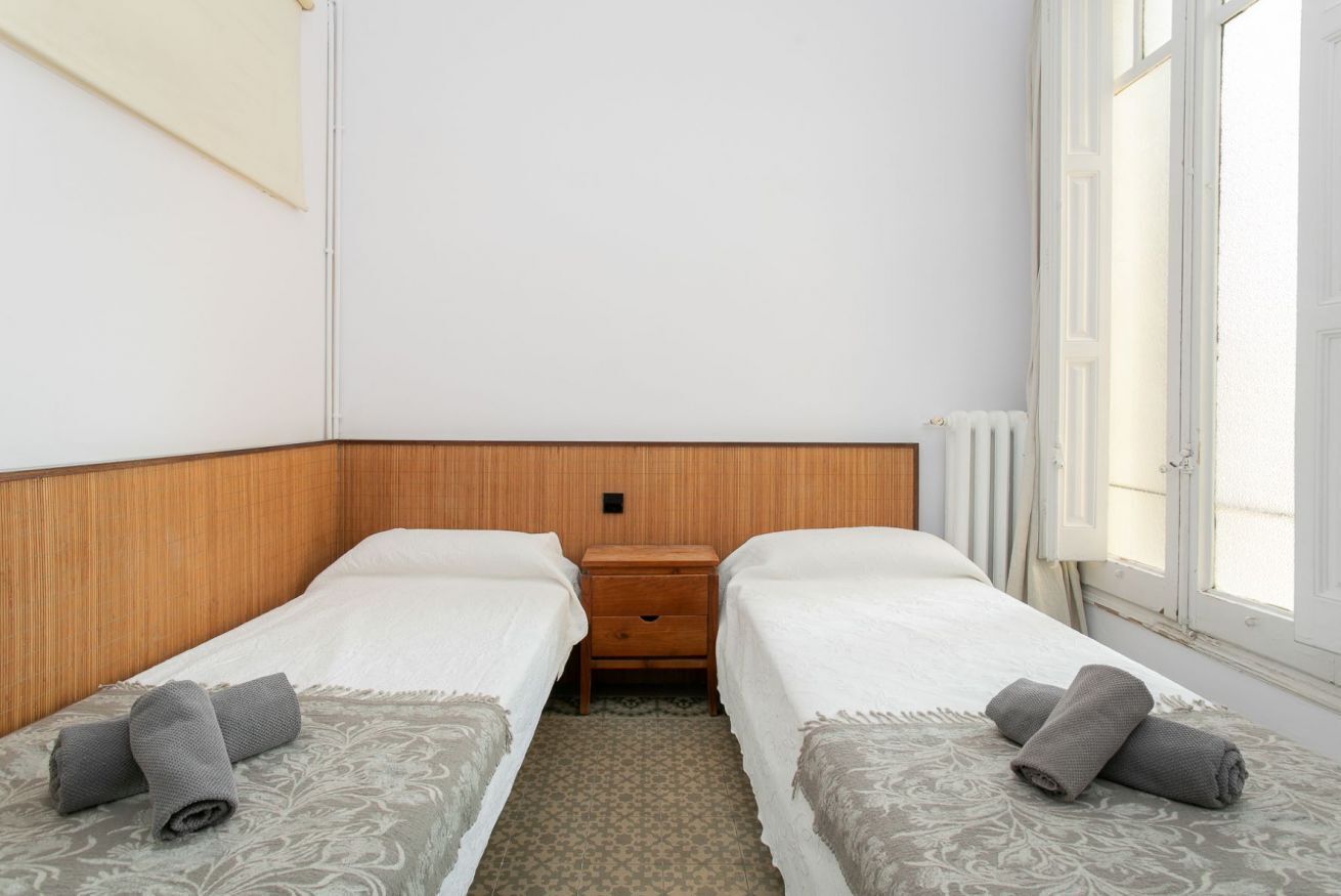 Habitación de hotel con dos camas, cabecera de madera, mesita de noche y ventana con cortinas.