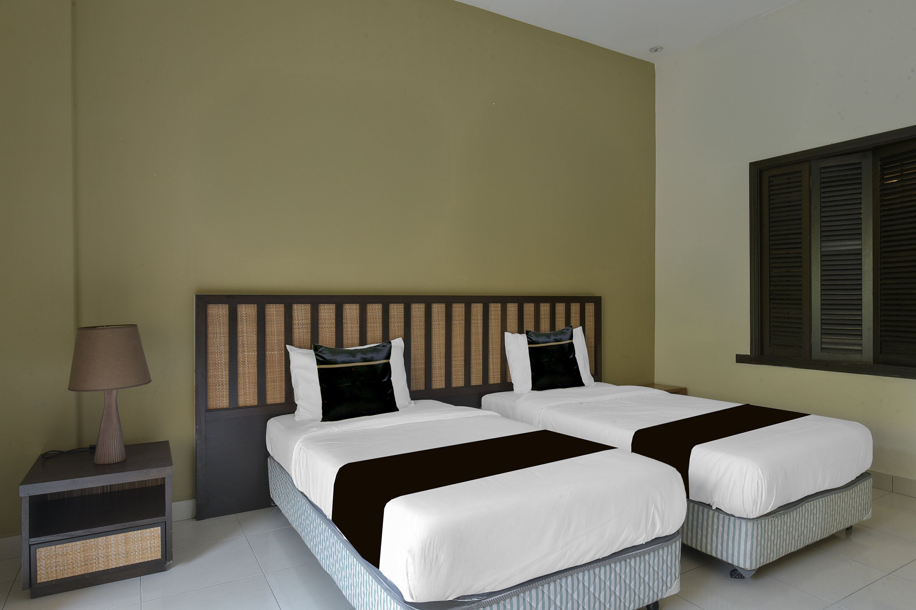 A hotel room with two beds, a nightstand with a lamp, and a window with shutters.