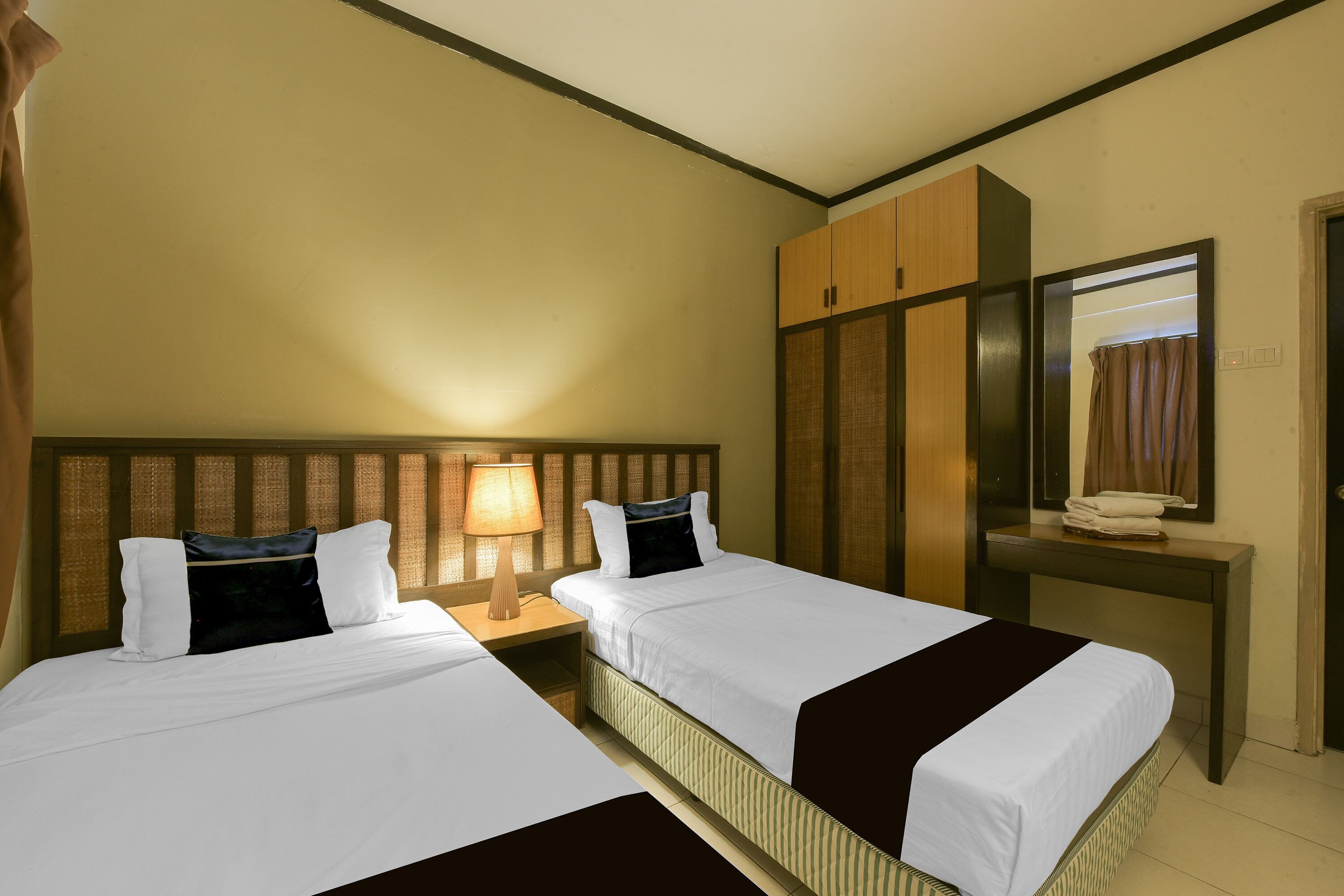 Habitación de hotel con dos camas, una mesita de noche con lámpara, un armario y un espejo.