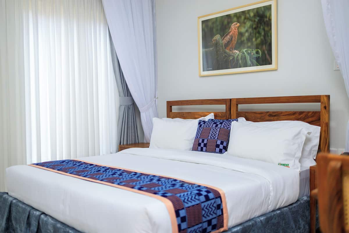 Una cama bien hecha con una colcha estampada, un cuadro en la pared y una ventana con cortinas traslúcidas.