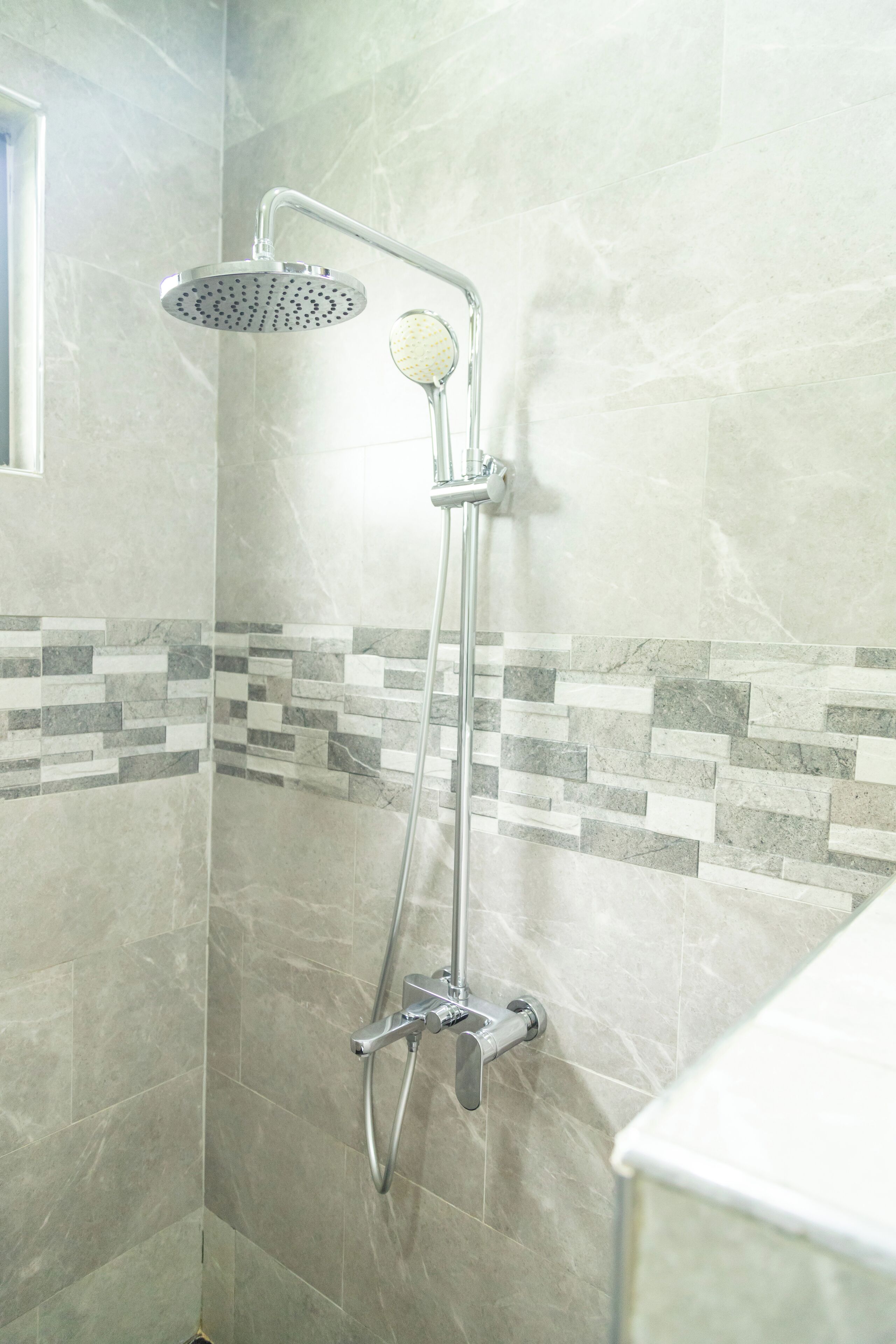 A shower with a handheld showerhead and a wall-mounted rain showerhead in a bathroom with tiled walls and a mosaic tile border.