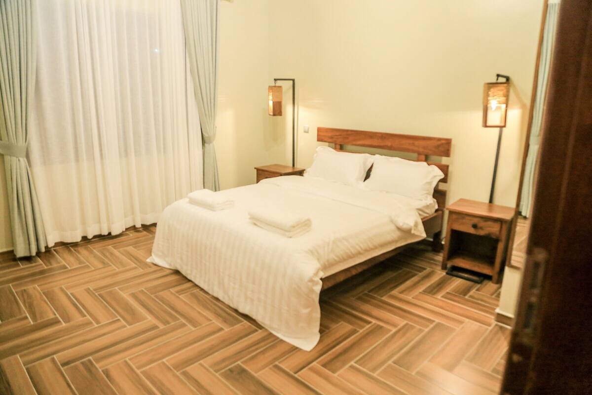 Una cama bien hecha con cabecera de madera, dos mesitas de noche con lámparas y un suelo de madera con patrón de espiga.