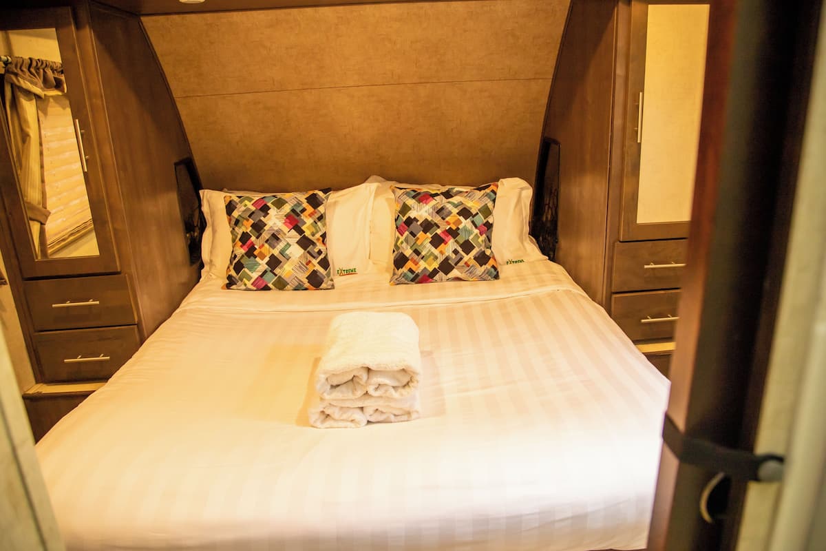 Una cama bien hecha con almohadas estampadas y una toalla doblada, cabecero de madera y una ventana pequeña con cortinas.