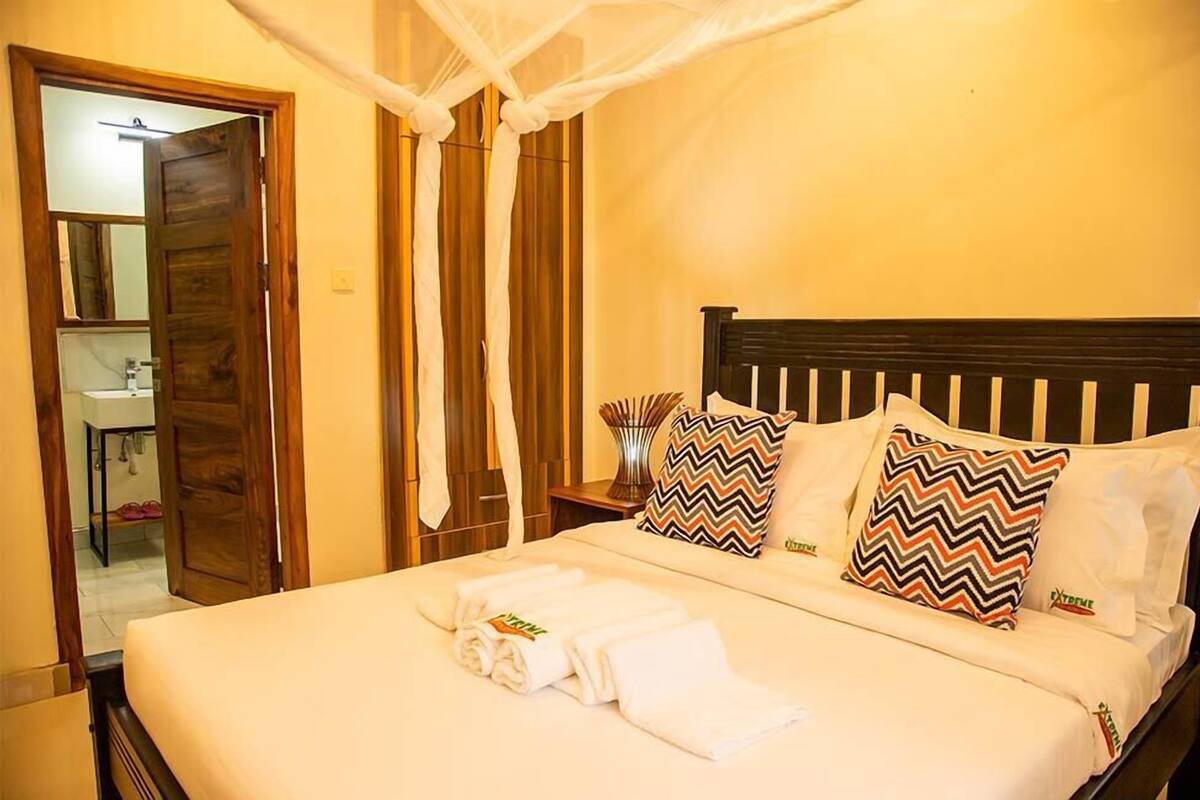 Una cama bien tendida con sábanas blancas y dos almohadones decorativos, un cabecero de madera y una mesita de noche con un florero.