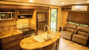 A compact kitchen with a stove, microwave, sink, and a refrigerator. There's a seating area with a couch and a window with blinds. - Extreme Adventure Park (Busika)