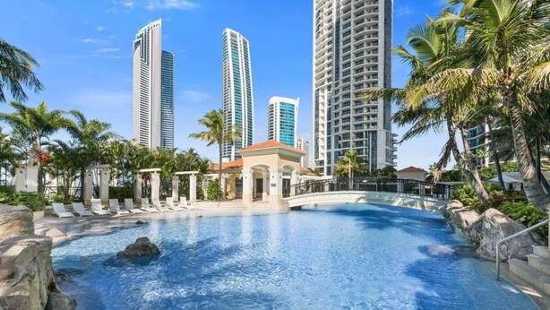 Uma área de piscina com espreguiçadeiras, um elemento decorativo de pedras e edifícios altos ao fundo.