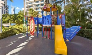 A playground with a yellow slide and climbing structures, surrounded by palm trees and a building in the background. - 26th Floor at Chevron (Surfers Paradise)