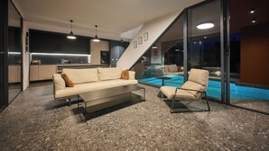 Ett modernt vardagsrum med en soffa och en fåtölj, en glasvägg med utsikt över en pool och ett kök i bakgrunden.