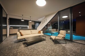 A modern living area with a sofa and armchair, a glass wall overlooking a pool, and a kitchen in the background. - 3-bedroom Villa Prima in Mugeba (Mugeba)