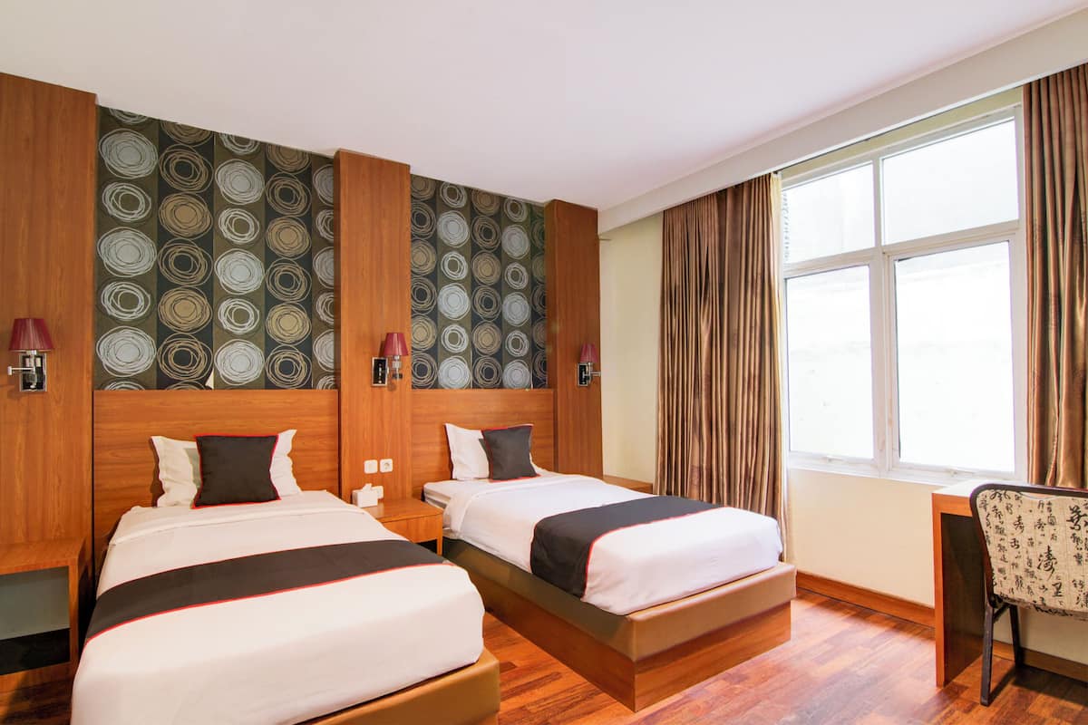Ein Hotelzimmer mit zwei Betten, Holz-Kopfteilen, einem großen Fenster mit Vorhängen und einem Schreibtisch mit Stuhl.