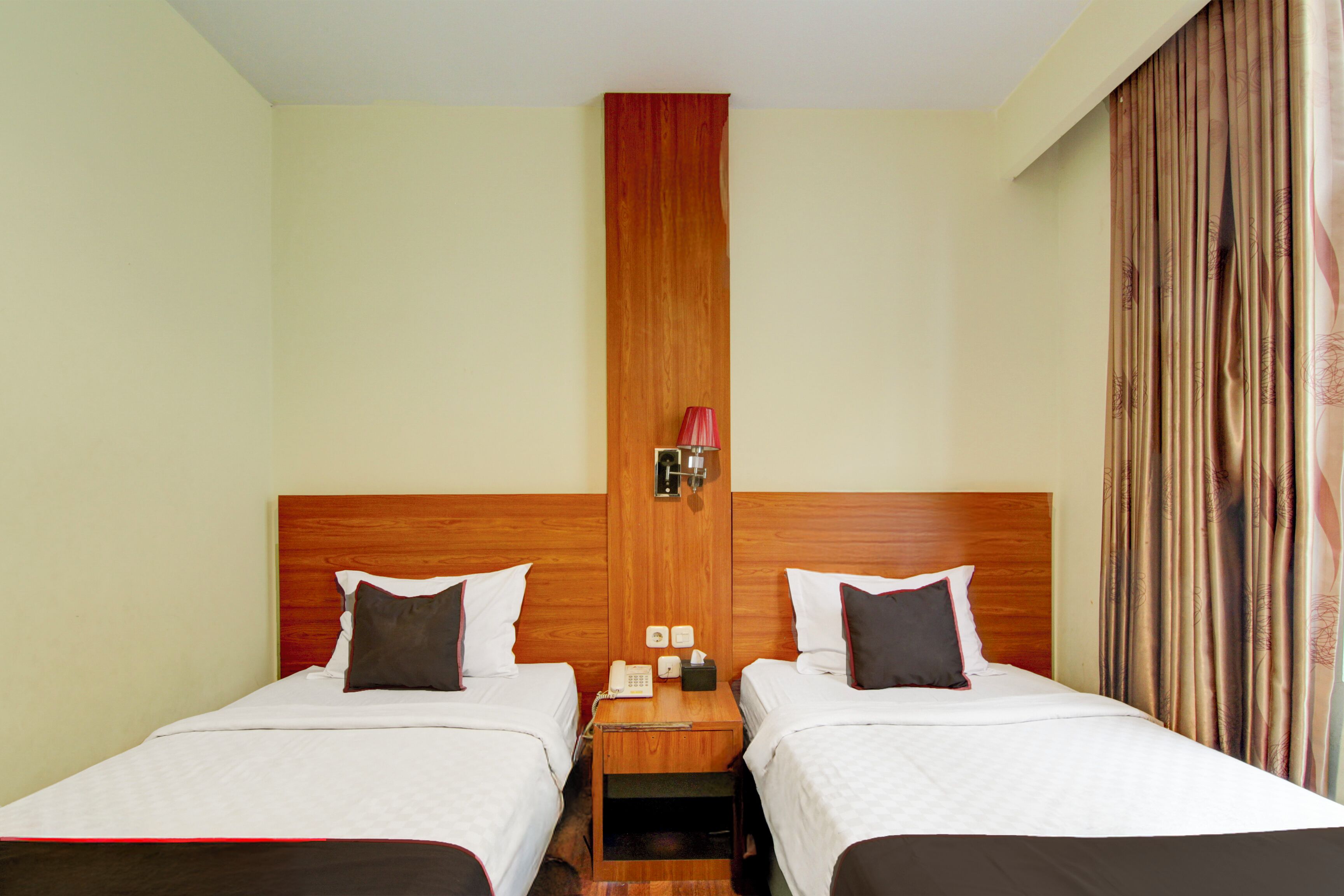 Ein Hotelzimmer mit zwei Betten, Holzkopfteilen, einem Nachttisch mit Telefon und Taschentüchern sowie einem Vorhang am Fenster.
