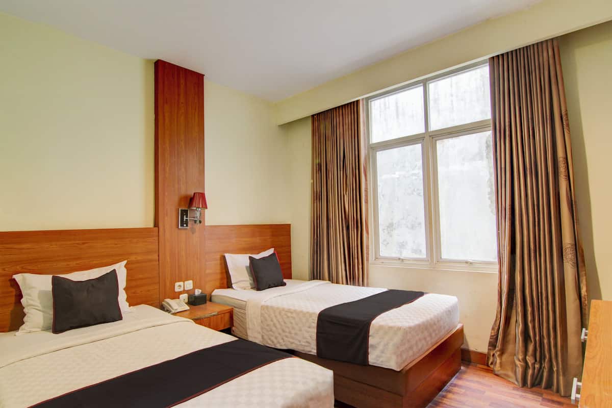 Ein Hotelzimmer mit zwei Betten, einem Holzkopfende, einem Fenster mit Vorhängen, einem Nachttisch mit Telefon und einer Wandleuchte.