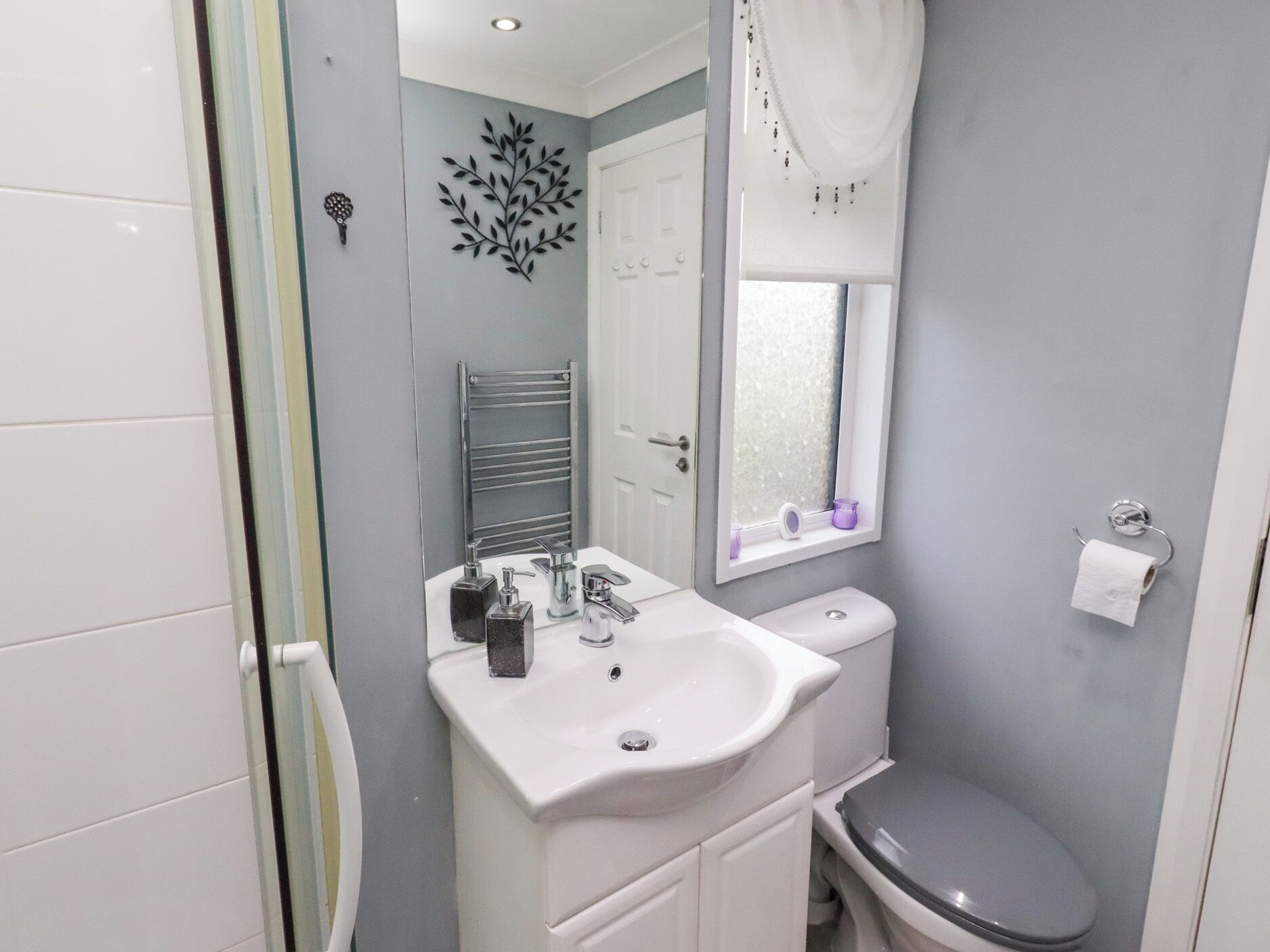 Un baño con lavamanos blanco, inodoro, espejo y ventana con cortina.