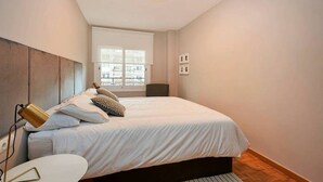 A bedroom with a bed, bedside table, lamp, window, and a view of buildings. - Design Flat, Terrace / Paseo de Gracia, Born (Barcelona)