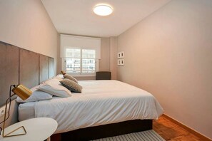 Ein Schlafzimmer mit Bett, Nachttisch, Lampe, Fenster und Blick auf Gebäude.