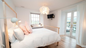 Ein Schlafzimmer mit einem hölzernen Bett, weißer Bettwäsche, einem an der Wand montierten Fernseher und einem Balkon mit Sitzgelegenheit.