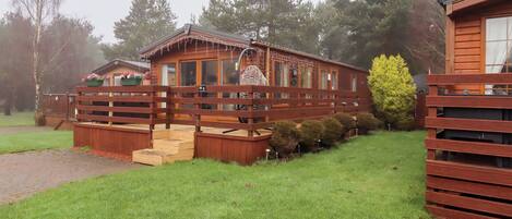 A wooden cabin with a deck, surrounded by a wooden fence and a grassy area.