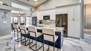 A modern kitchen with white cabinets, a large island with bar stools, and a stainless steel refrigerator. - Luxe Waterfront Cape Coral Home w/ Gulf Access! (Cape Coral)