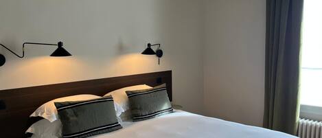 A neatly made bed with white linens and two pillows, a wooden headboard, and two wall-mounted reading lamps.
