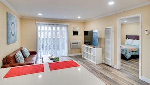 A modern living room with a brown leather sofa, a white coffee table, and a red rug. There is a TV on a white stand, a white shelving unit, and a bed with a blue floral bedspread in the adjacent room. - 2 Mi to Dtwn Pleasanton Condo w/ Pool Access (Pleasanton)