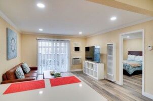 Una sala moderna con un sofá de cuero marrón, una mesa de centro blanca y una alfombra roja. Hay un televisor sobre un mueble blanco, una estantería blanca, y una cama con colcha estampada de flores azules en la habitación contigua.
