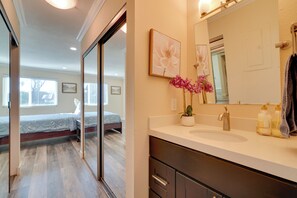 Een slaapkamer met een bed en een badkamer met een wastafel, spiegel en een bloevas.