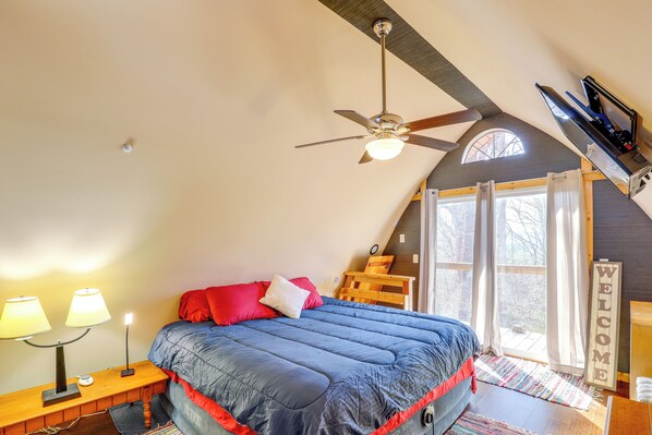 A bedroom with a sloped ceiling, a bed with a blue comforter, two bedside lamps, a wooden nightstand, a ceiling fan, and a window with curtains. - 1 Mi to Tiec: Peaceful Mountain Cabin (Mill Spring)