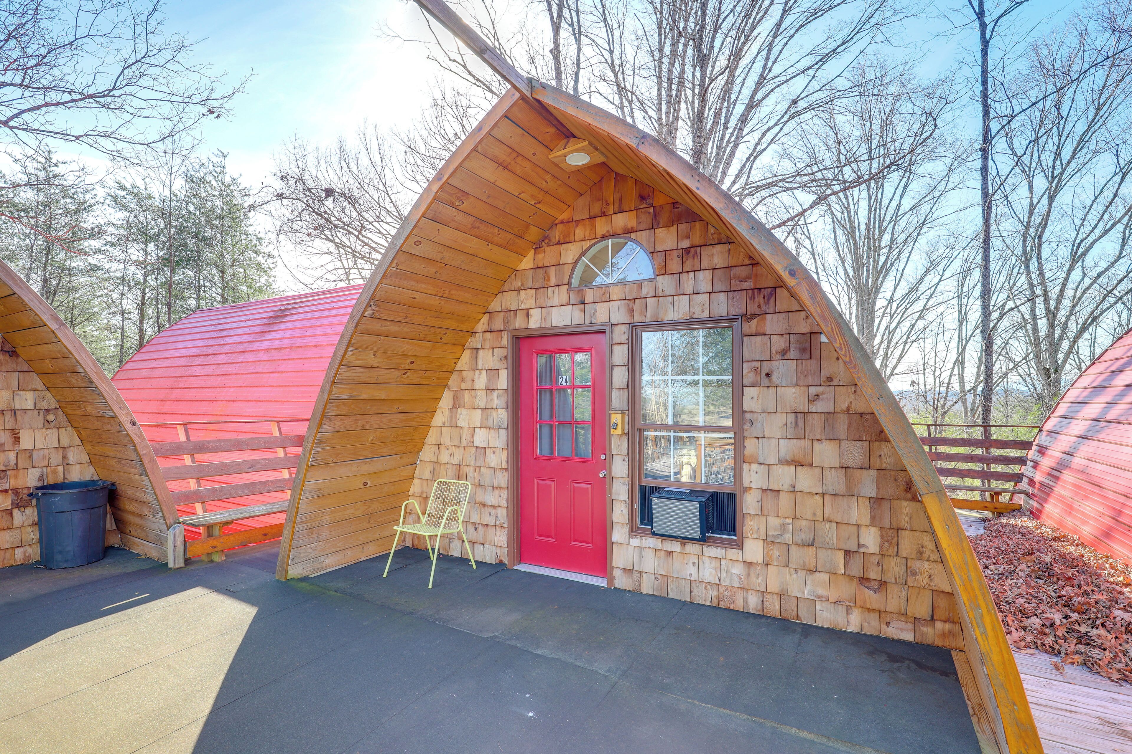 A wooden A-frame structure with a red door and a window, surrounded by trees.