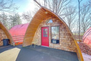 Une structure en bois en forme de toit à deux versants, avec une porte rouge et une fenêtre, entourée d’arbres.